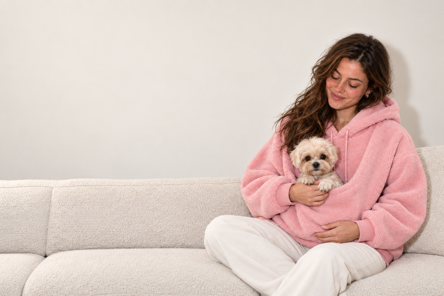 Woman seated on a cream sofa wearing a pink Pawttle hoodie, supporting a small dog in the front pouch.